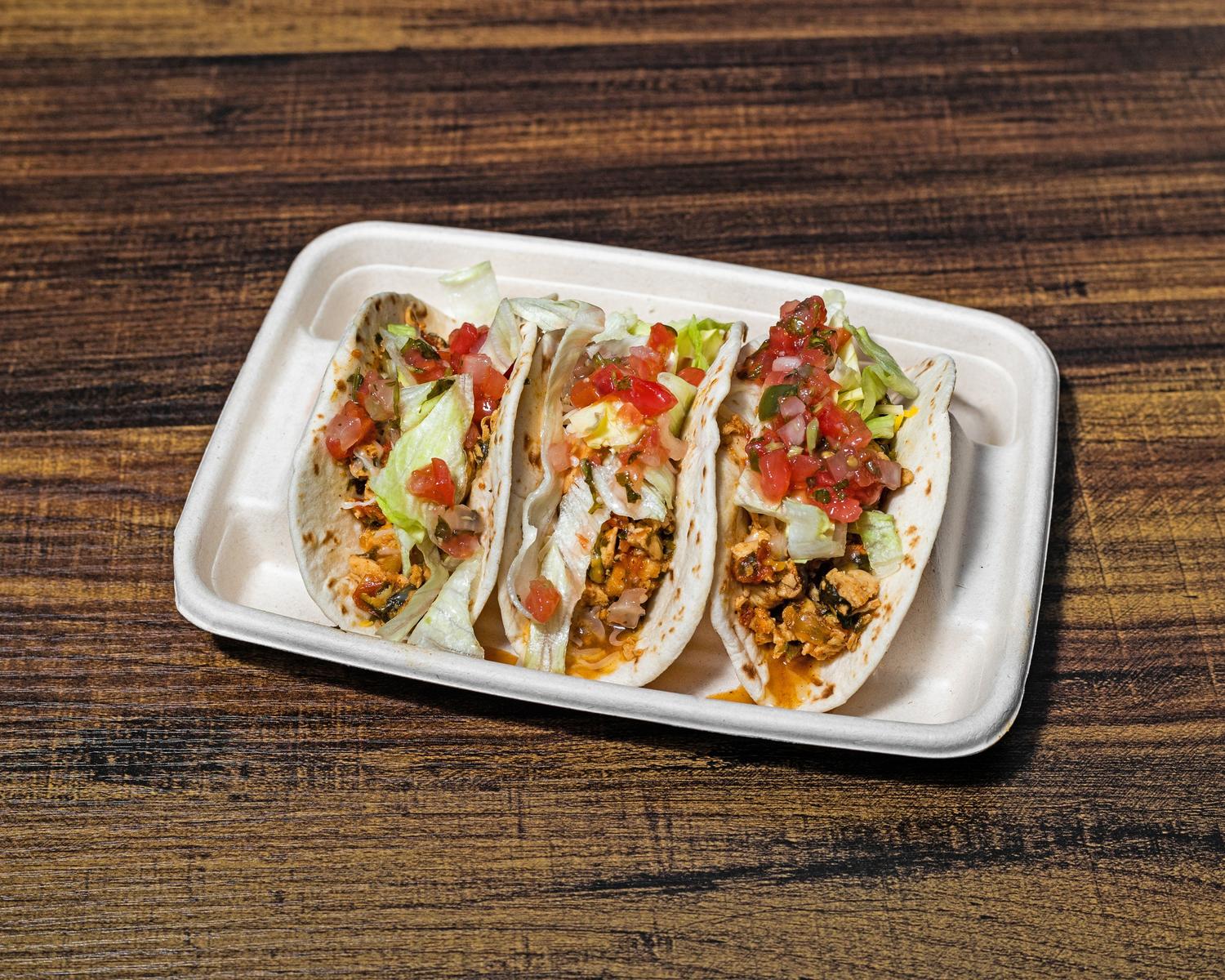Three tacos topped with lettuce, cheese and salsa in a cardboard tray